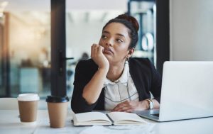 Woman Showing Signs of Burnout at Work Unhappily Staring into the Distance at Office Desk with Laptop and Coffee Cups | Vitacost.com/blog
