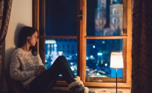 Woman Struggling With Winter Sleep Sitting on Bench in Bedroom Starting Blankly Out Window into Bustling City Street | Vitacost.com/blog