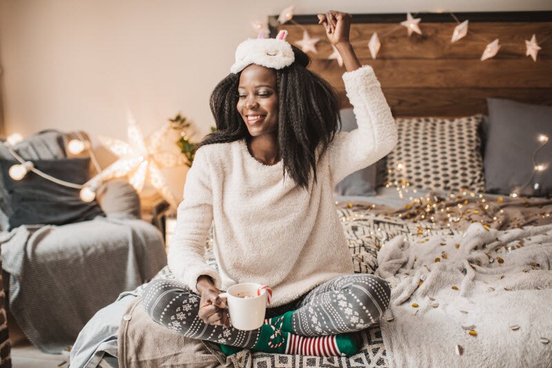 How to Be Mindful This Holiday Season Woman Who Learned How to Be Mindful During the Holidays Sitting Happily on Bed With Mug of Hot Cocoa in Hand | Vitacost.com/blog