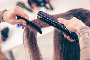 Stylist Using Flat Ironing Tool on Woman's Long Brown Hair to Represent Concept of How to Protect Hair From Heat Damage | Vitacost.com/blog