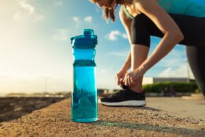 Concept of How to Go Green in Your Exercise Routine Represented by Athletic Woman Tying Shoe Beside Reusable Blue Water Bottle on Gravel Surface | Vitacost.com/blog