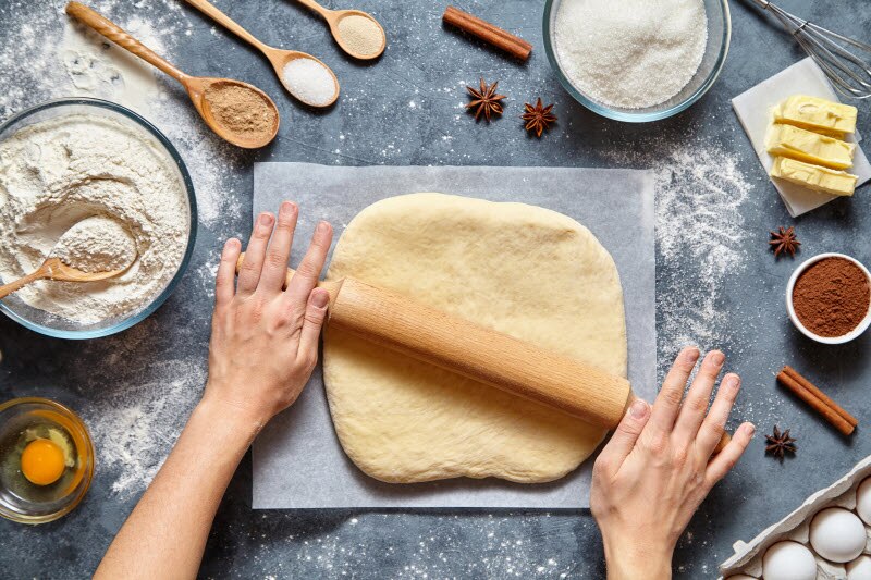 Overhead View of Dough Being Rolled Out on Parchment Paper Surrounding by Ingredients in Bowls and Spoons to Demonstrate How to Make a Pie | Vitacost.com/blog