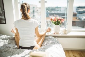 Ponytailed Woman on Bed Facing Window Meditating After Learning to Relieve Anxiety Naturally With Deep Breathing Techniques | Vitacost.com/blog