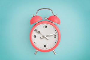Concept of Intermittent Fasting for Beginners Represented by Coral-Colored Alarm Clock with Fork & Knife for Hands Against Solid Light Teal Background | Vitacost.com/blog
