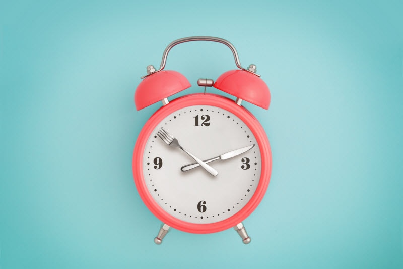 Concept of Intermittent Fasting for Beginners Represented by Coral-Colored Alarm Clock with Fork & Knife for Hands Against Solid Light Teal Background | Vitacost.com/blog