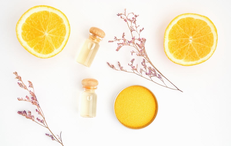 Vitamin C Benefits for Skin Represented by Overhead Shot of Oranges, Two Canisters of Serum, Orange Scrub and Flowering Twigs on White Surface | Vitacost.com/blog