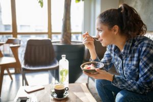 Woman Sitting on Couch Beside Coffee Table Eating Bowl Full of Fresh Fruit and Cereal as Example of Foods Not to Eat Before Bedtime | VItacost.com/blog