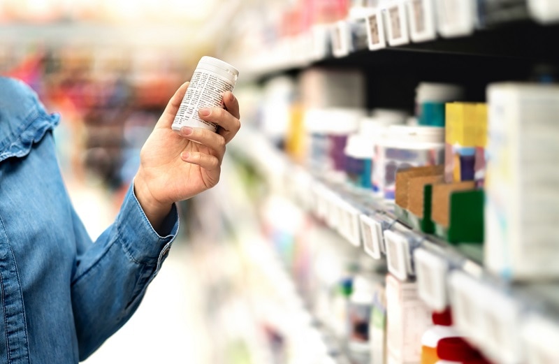 Woman Following Tips on How to Choose a Probiotic Supplement Reading Bottle Label Standing Beside Store Shelves | Vitacost/com/blog