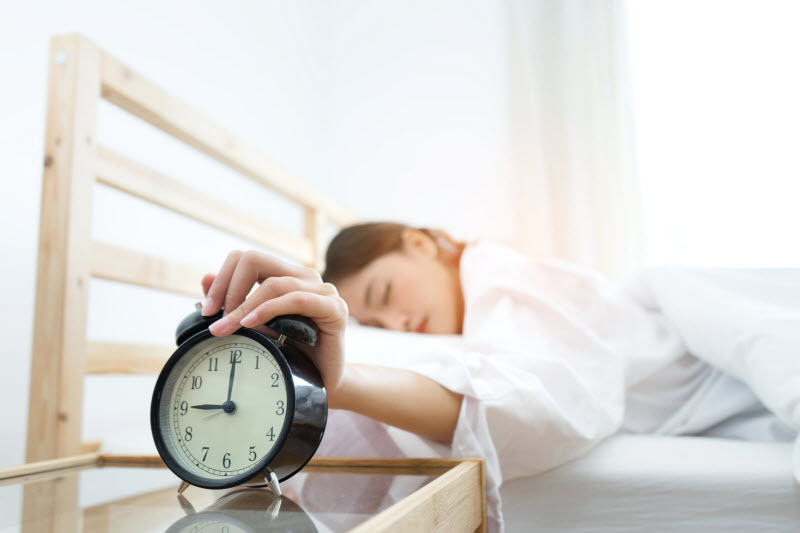 Woman Following Bedtime Routine in Bed Hitting Snooze Button on Alarm Clock on Bedside Table | Vitacost.com/blog