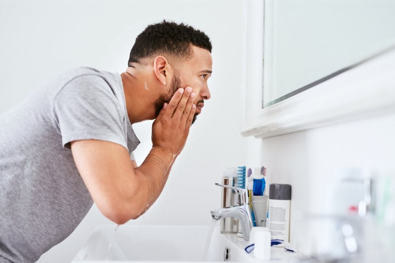 Side View of Guy with Beard Applying the Best Skincare Products for Men to His Face in Front of Bathroom Mirror | Vitacost.com/blog