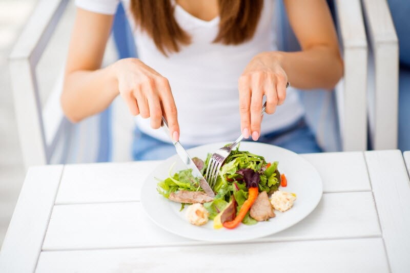 Torso View of Woman Learning How to Eat for Your Blood Type Enjoying Salad Sitting on White Wood Chair at White Wood Table | Vitacost.com/blog