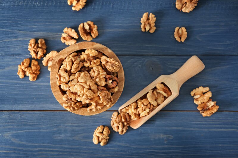 How to Enjoy the Many Health Benefits of Walnuts The Health Benefits of Walnuts Represented by Wooden Bowl and Scoop Filled With Walnut Halves on Dark Blue Wooden Surface | Vitacost.com/blog