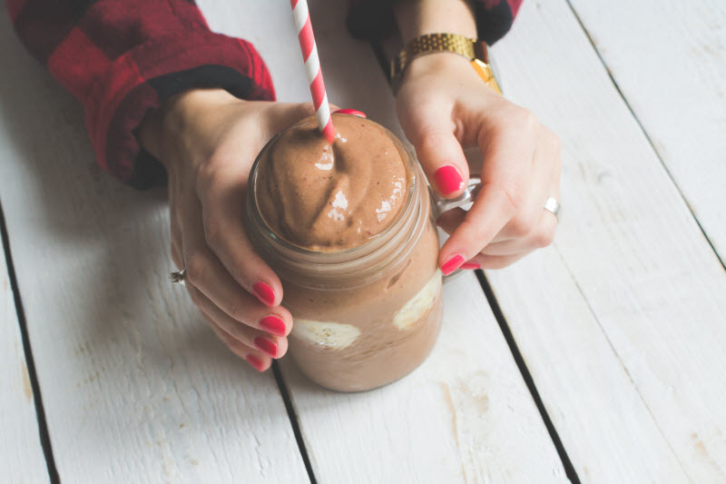 Overhead View of Chocolate Protein Powder Mixed into a Smoothie in a Jar with Red & White Straw Held by Woman | Vitacost.com/blog