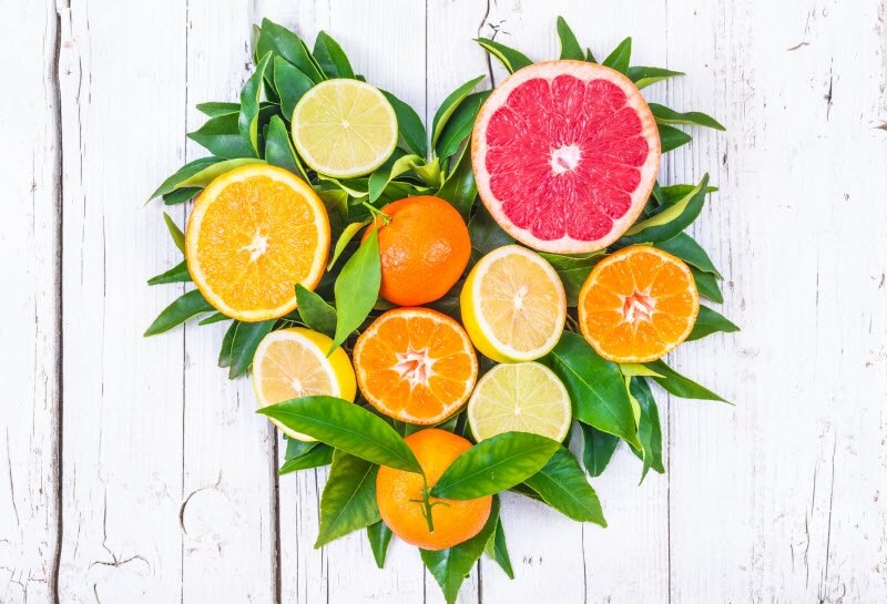 Benefits of Citrus Fruits Represented by Variety of Halved Oranges, Lemons and Grapefruit in Heart Shape with Green Leaves on White Wood Surface | Vitacost.com/blog