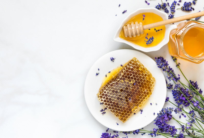 Different Types of Honey Represented by Plates & Jar Filled with Honey and Honeycomb Surrounded by Lavender Flowers on White Marble Surface | Vitacost.com/blog