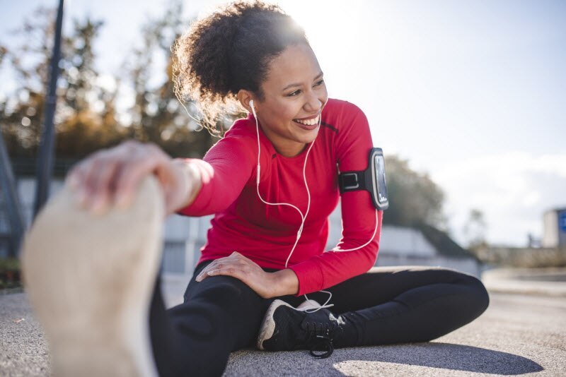 Woman With Headphones in Red Workout Shirt and Black Pants Stretching on Pavement as Part of Plan for Getting in Shape at 40 | Vitacost.com/blog