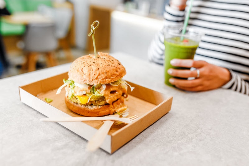 Torso View of Woman Leaning How to Eat Healthy at a Restaurant Holding Green Smoothie at Table with Veggie Burger on Tray | Vitacost.com/blog