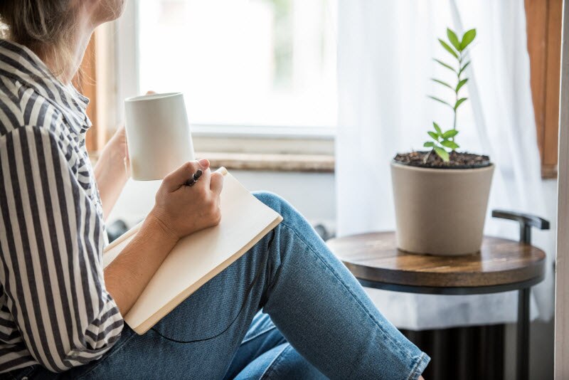 Woman Following Tips on How to Stay Focused Sitting Beside Window Writing in Notebook With Mug of Coffee in Hand | Vitacost.com/blog