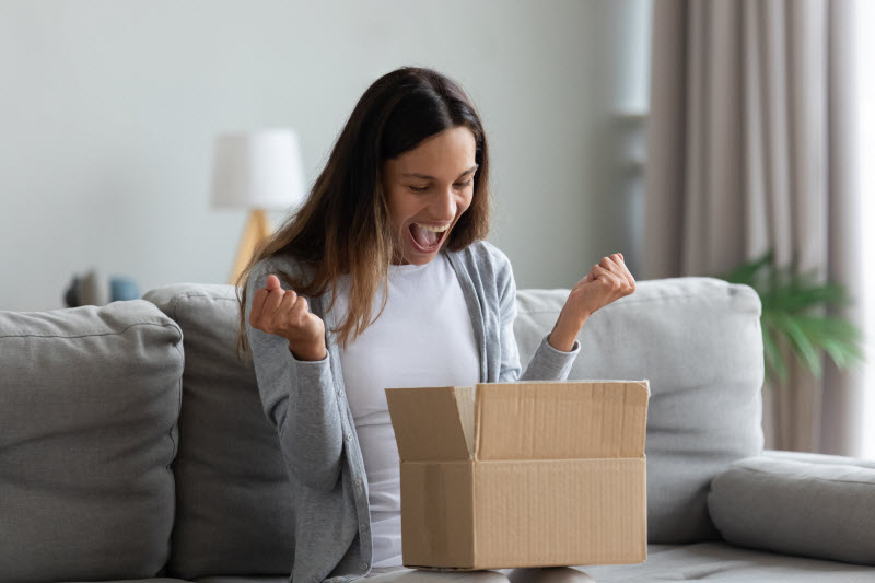 Elated Woman Sitting on Couch Opening Box of Essentials for Healthy Living From Vitacost | Vitacost.com/blog