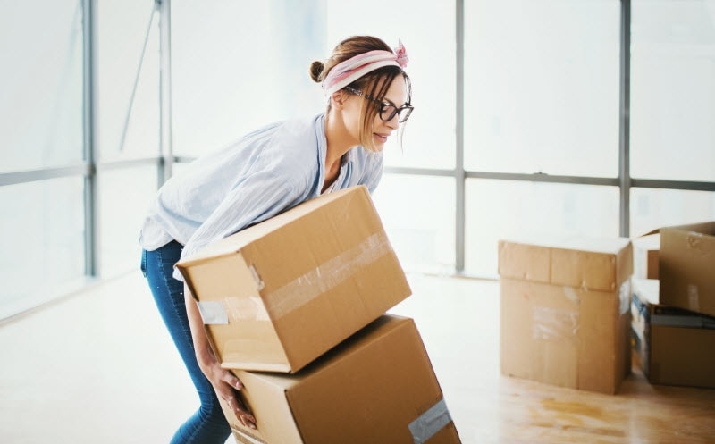 Woman Following Garage Cleaning Tips Moving Stack of Boxes to Organize Her Cluttered Garage | Vitacost.com/blog