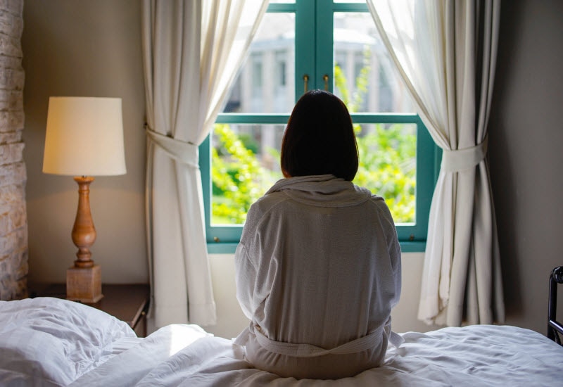 Back View of Woman Wondering How to Sleep When You Have Anxiety Sitting in Bed Looking Out Window | Vitacost.com/blog