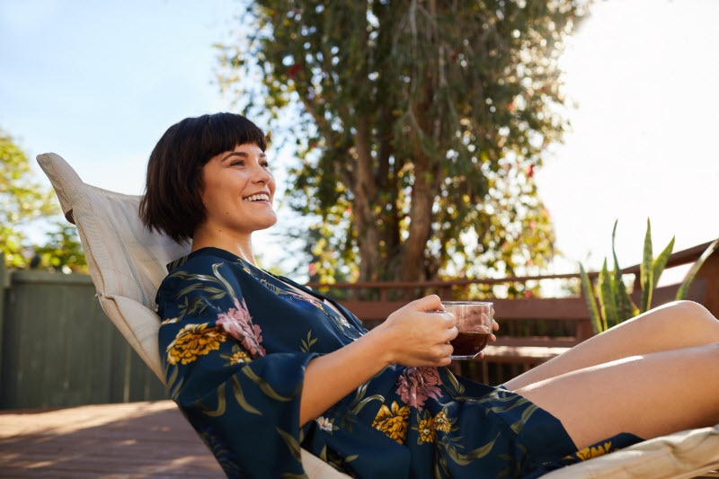 Smiling Woman Holding Cup of Tea Relaxing on Patio Chair While Taking Mental Health Day | Vitacost.com/blog