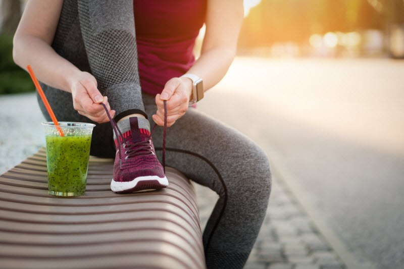 Woman Following Preworkout Fitness Advice Tying Shoe on Park Bench with Green Smoothie at Side | Vitacost.com/blog