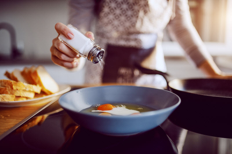 Woman Not Watching Salt Intake Shaking Salt into Bowl Bowl of Eggs on Kitchen Counter | Vitacost.com/blog
