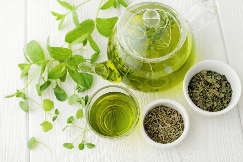 Clear Glass Pot and Mug Filled With Top Detox Teas With Herbs in Bowls and Greenery on White Wood Surface | Vitacost.com/blog