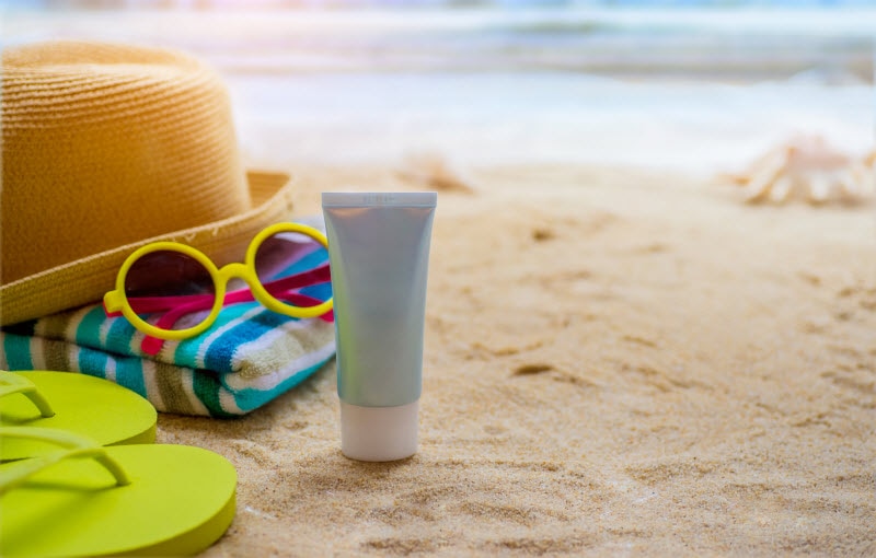Concept of What is Sunscreen Represented by Beach Scene with Sunscreen, Towel, Hat and Glasses on Sand | Vitacost.com.blog