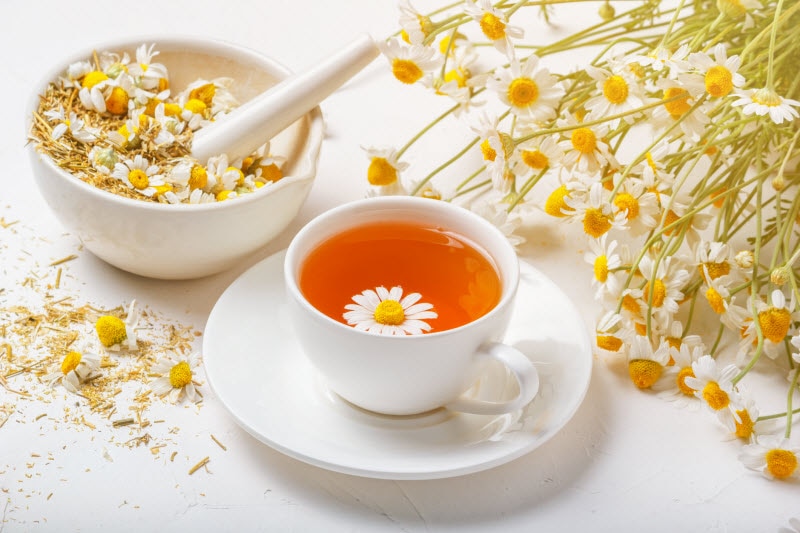 Mug of Chamomile Tea with Bunch of Chamomile Flowers and Bowl of Dried Petals to Represent Chamomile Benefits | Vitacost.com/blog