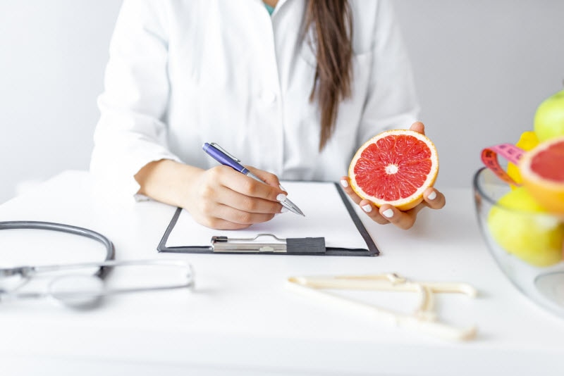 Concept of What Does a Dietitian Do Represented by RD in Lab Coat Writing on Clipboard While Holding Grapefruit | Vitacost.com/blog