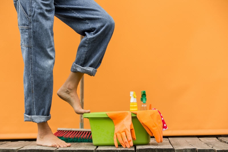 Natural House Cleaning Tips for Fall Woman in Loose Jeans Propping Foot on Bucket of Eco Friendly Cleaning Supplies Against Orange Background | Vitacost.com/blog