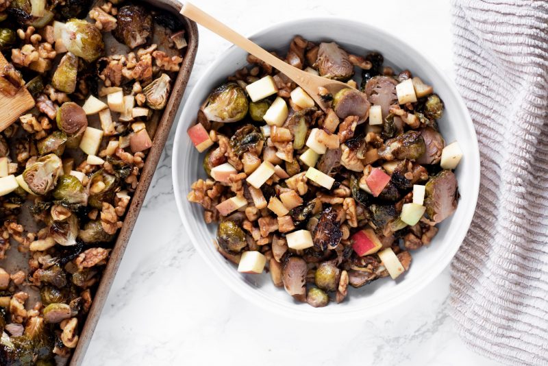 Top View Roasted Brussels Sprouts Apple and Walnuts in Bowl with Wooden Spoon Next to Sheet Pan | Vitacost Blog