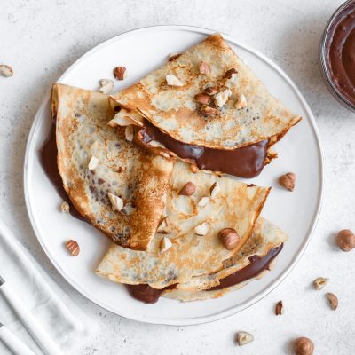 Three Crepes Folded into Triangles and Filled with Chocolate Sauce on White Plate Garnished with Hazelnuts | Vitacost Blog