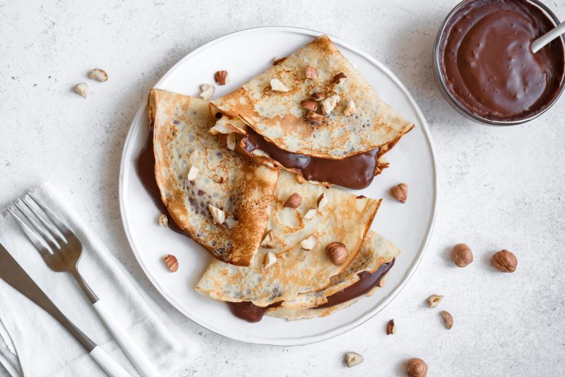 Three Crepes Folded into Triangles and Filled with Chocolate Sauce on White Plate Garnished with Hazelnuts | Vitacost Blog