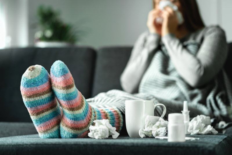 6 Weak Immune System Symptoms + Solutions Woman With Weak Immune System Symptoms Blowing Nose on Couch With Feet Up Surrounded by Tissues and Cold Remedies | Vitacost.com/blog