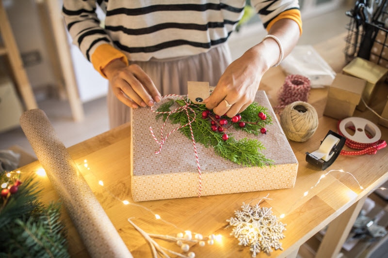Torso View of Woman Wrapping Gifts Under $30 on Table with Holiday Lights, Greenery & Gift Wrap | Vitacost.com/blog