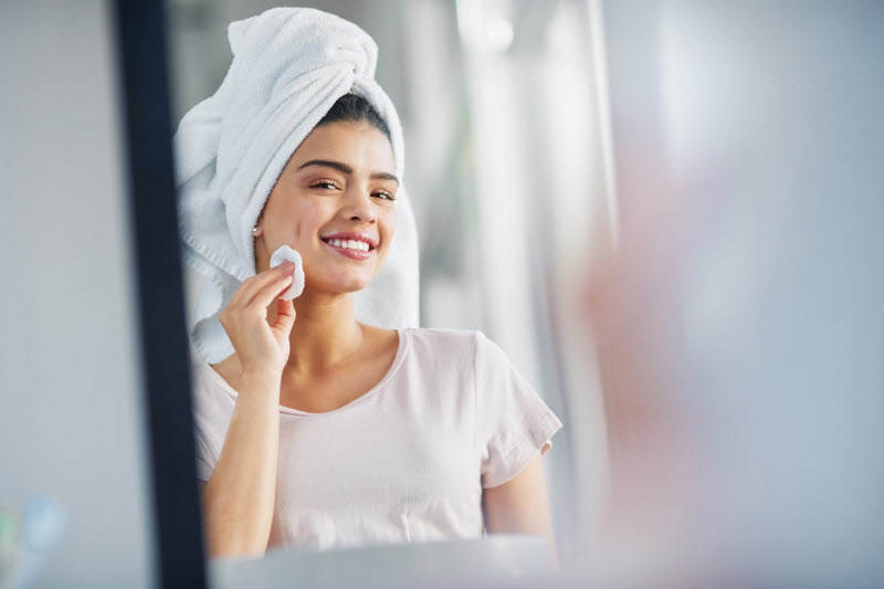 Woman in Mirror Following Exfoliating For Beginners Tips Wiping Product on Face with Cotton Pad | Vitacost.com/blog