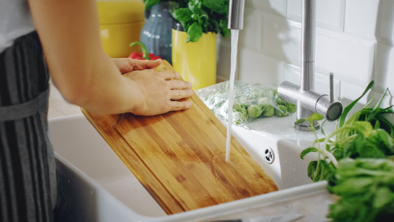 Person Who Learned How to Clean a Wooden Cutting Board Scrubbing Board Under Running Water in Sink | Vitacost.com/blog
