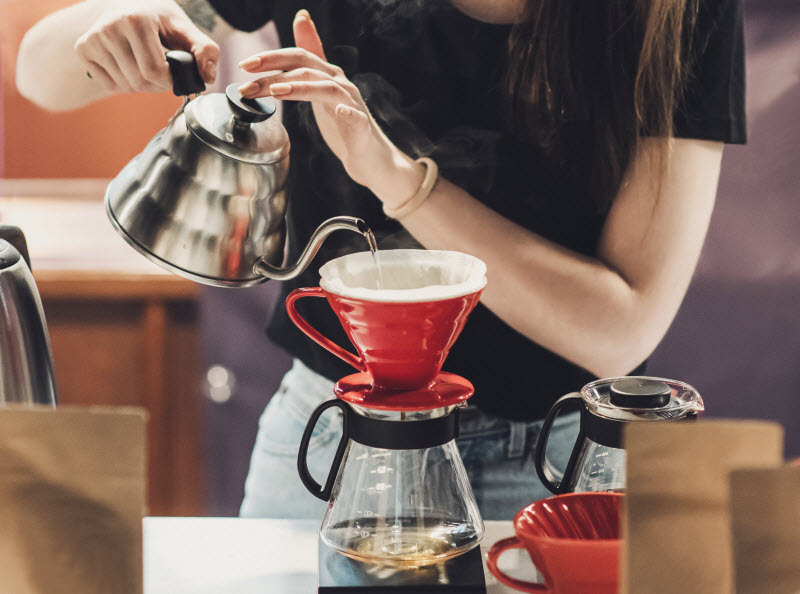 Woman Who Learned How to Make Pour Over Coffee Pouring Hot Water into Red Coffee Filter Funnel | Vitacost.com/blog