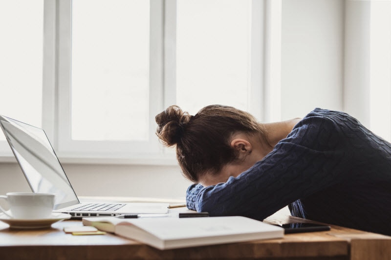 Woman at Desk with Laptop Resting Head on Arms Exhausted Due to Vitamin B12 Deficiency | Vitacost.com/blog