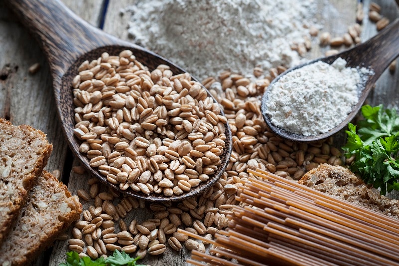 Overhead View of Spelt Grains and Spelt Flour on Spoons alongside Spelt Pasta and Spelt Bread | Vitacost.com/Blog