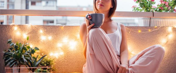 Woman Enjoying Cup of Self Care Essentials Tea on Balcony With Plants and String Lights | Vitacost.com/blog