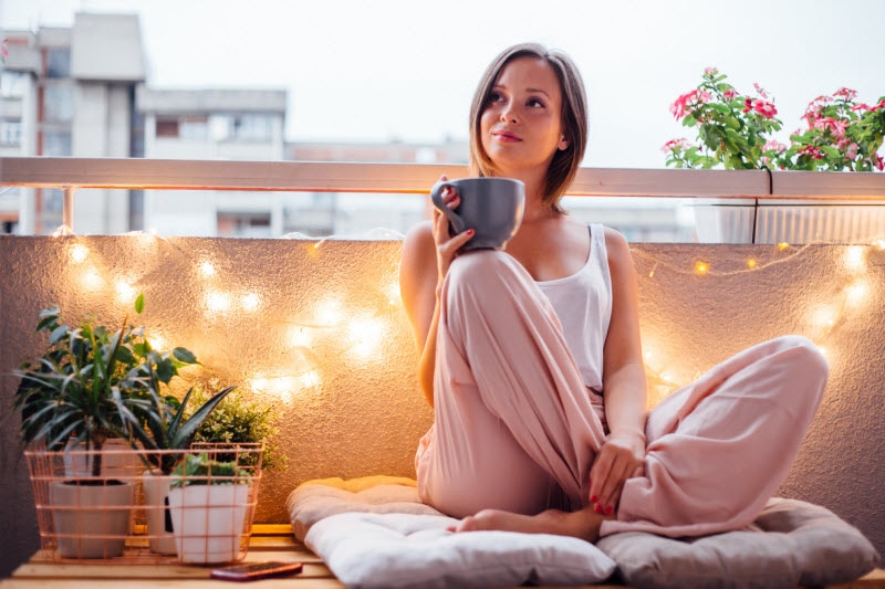 Woman Enjoying Cup of Self Care Essentials Tea on Balcony With Plants and String Lights | Vitacost.com/blog