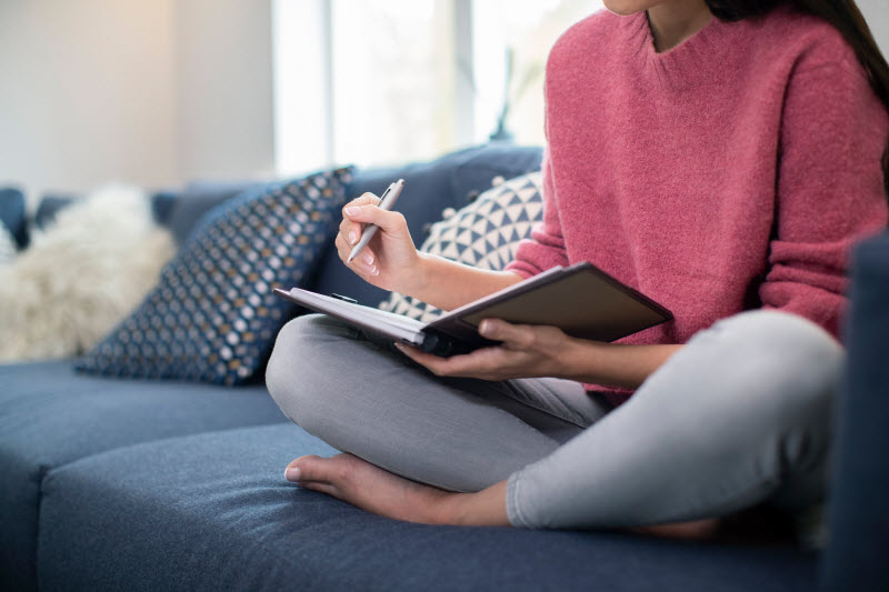 Woman Learning How to Put Yourself First Sitting on Couch Writing Self-Care Checklist in Journal | Vitacost.com/blog