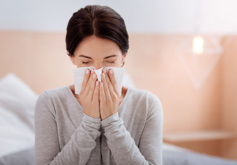 Woman in Gray Shirt Learning What are Antibodies Sneezing into Tissue | Vitacost.com/blog
