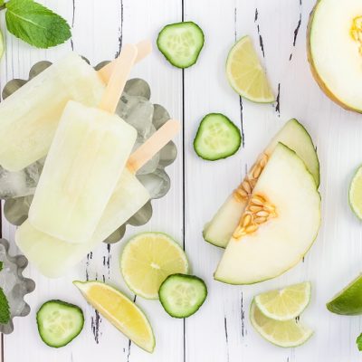 Light Yellow Popsicles Surrounded by Sliced Cucumbers, Lime Wedges, Honeydew and Sprigs of Basil on a White Wooden Background | Vitacost.com/blog
