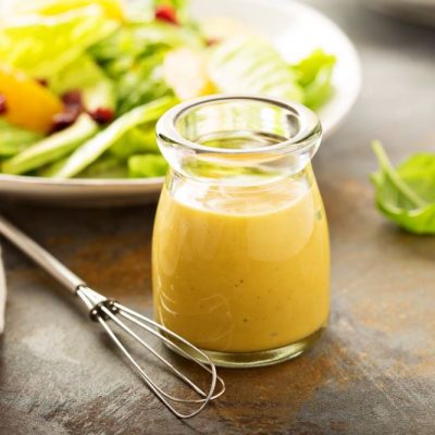 Honey Lemon Olive Oil Dressing in Glass Carafe on Wooden Table Next to Salad | Vitacost.com/blog