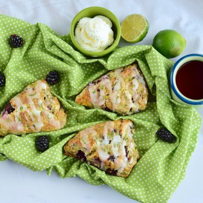 Three Blackberry Scones with Glaze on Green Cloth Surrounded by Blackberries, Cut Lime, Tea and Cream to Represent Blackberry Scone Recipe | Vitacost.com/Blog